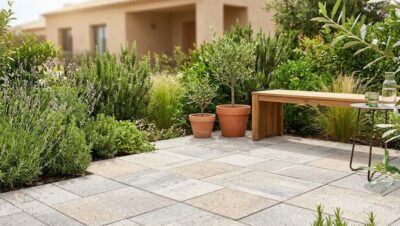 Pavimento de adoquín ecológico drenante en un patio residencial con plantas mediterráneas y mobiliario de madera. Solución sostenible para evitar charcos en el jardín en 2026.